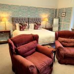 Two wingback red chairs in front of a superking bed backed by an arts and crafts style wallpaper